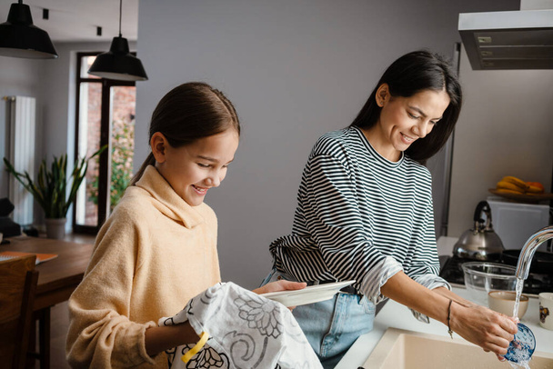Happy beautiful mother and daughter smiling and washing dishes in cozy kitchen - Fotografie, Obrázek