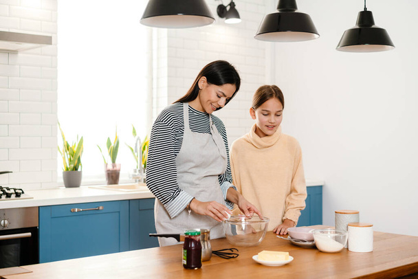 Happy beautiful mother and daughter smiling and making pancake dough in cozy kitchen - Foto, Bild