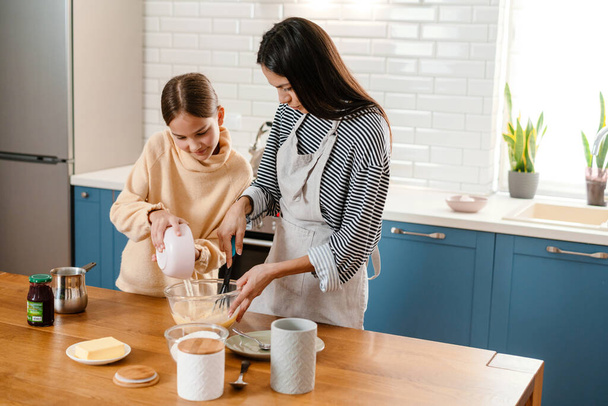 Happy beautiful mother and daughter smiling while making pancake dough in cozy kitchen - Foto, immagini