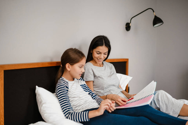 Happy mother and daughter doing homework while laying on bed indoors - Фото, зображення