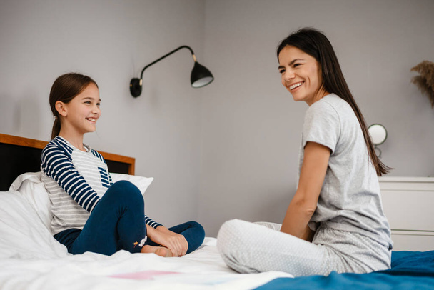 Joyful beautiful mother and daughter laughing while sitting on bed at home - Fotografie, Obrázek
