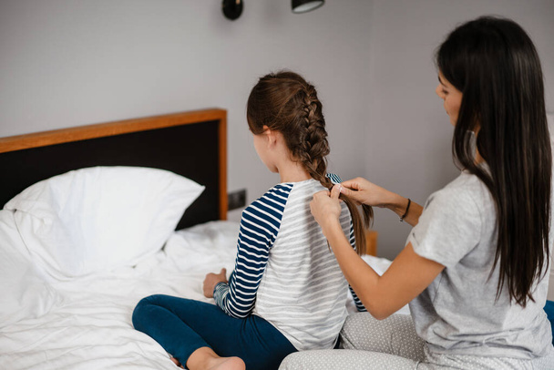 Nice beautiful mother making hairstyle to her daughter while sitting on bed at home - Foto, Imagen
