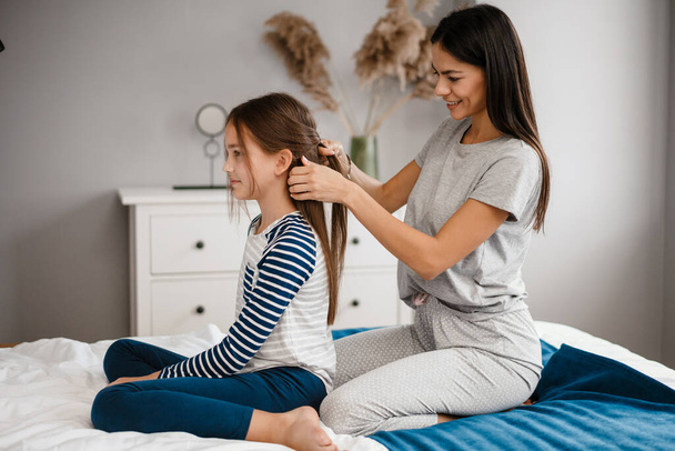 Beautiful young mother combing her cute little daughters hair while sitting on bed and smiling - Foto, afbeelding