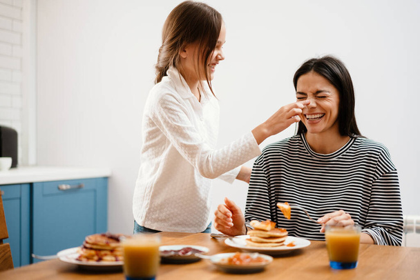 Cheerful beautiful mother and daughter making fun while having breakfast in cozy kitchen - Фото, изображение