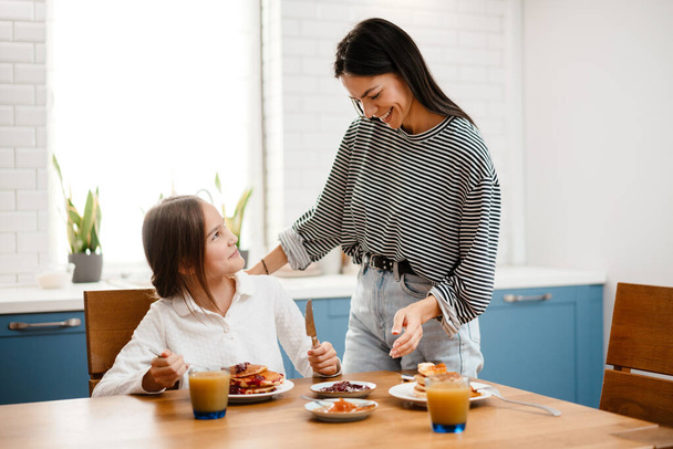 Cheerful beautiful mother and daughter smiling while having breakfast in cozy kitchen - Foto, imagen
