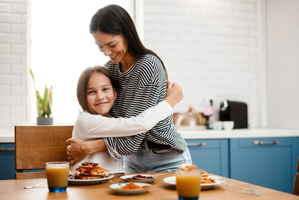 Cheerful beautiful mother and daughter hugging while having breakfast in cozy kitchen - Фото, изображение