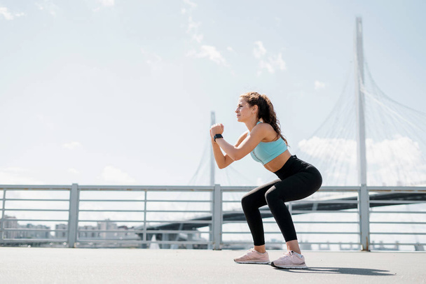A strong female fitness trainer does strength exercises squats outdoors. Healthy lifestyle concept. Slim figure and comfortable shoes. - Fotó, kép