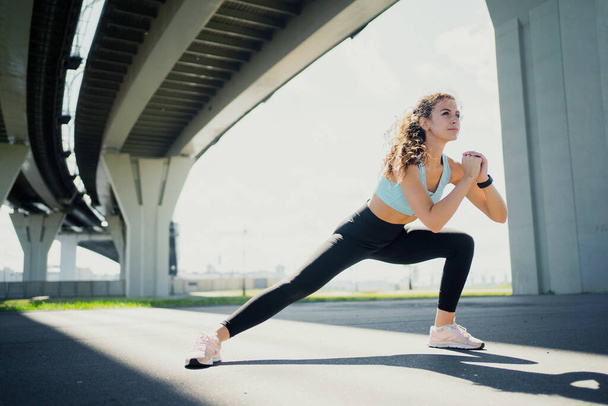 The athlete is a runner who leads a healthy lifestyle. Athletic figure a woman trains on the street in the city. Burns calories in the body. Exercises stretching muscles for fitness are active. - Foto, imagen