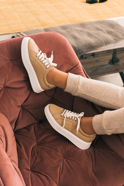 photo of a women legs in beige sports suit and stylish leather sneakers posing in the chair. selective focus - Photo, Image