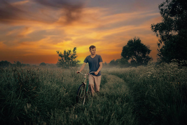 young man walks with a retro bicycle along a field road at sunset, an art photo of a lonely man. - Photo, Image
