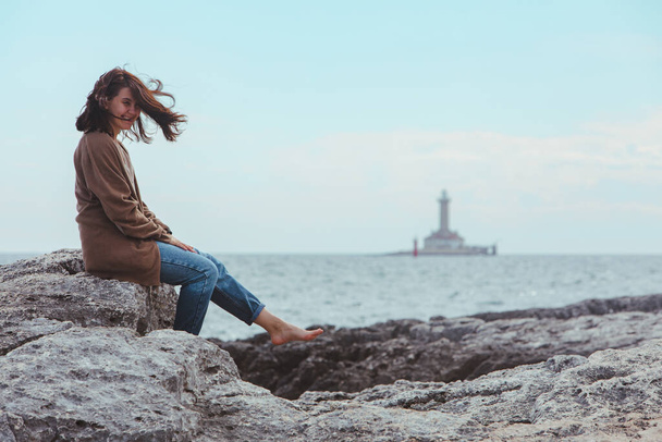 vrouw zittend door rotsachtige zee strand in natte jeans vuurtoren op de achtergrond. winderig weer. Zomervakantie. zorgeloos concept - Foto, afbeelding