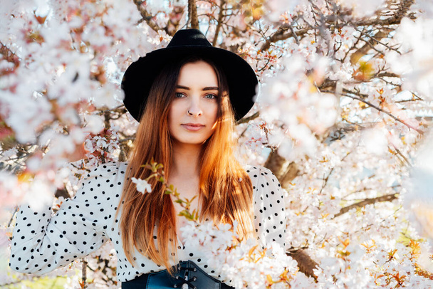 Young brunette woman in hat and dress in white pink blooming tree. Generation Z girl enjoy spring mood during sunny day. Springtime blossom. Selective focus, copy space - Photo, Image