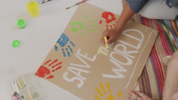 Alto angolo di mani di due giovani che lavorano segno slogan attivista dicendo salvare il mondo per il cambiamento climatico raduno, utilizzando gesso, seduto sul pavimento nella stanza dormitorio durante il giorno - Filmati, video