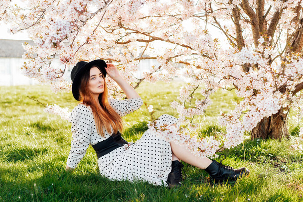 Young brunette woman in hat and dress sitting near white pink blooming tree. Generation Z girl enjoy spring mood during a sunny day. Relax on the nature. Springtime blossom. Selective focus. - Фото, зображення