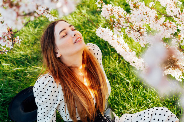 Young brunette woman in dress with closed eyes sitting near a pink blooming tree and enjoying the sunshine. Generation Z girl enjoy spring mood during a sunny day. Relax on the nature. - Fotó, kép