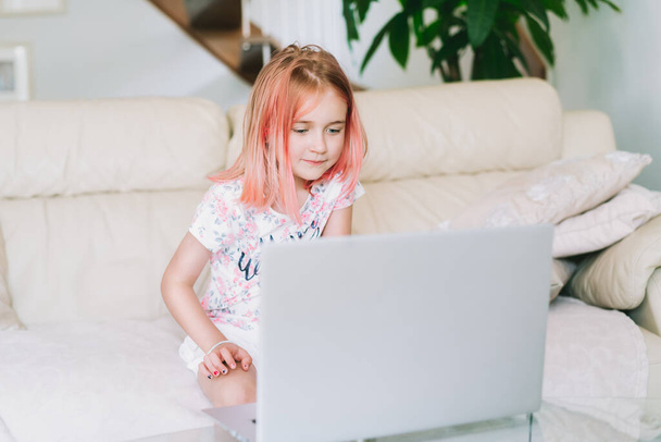 Kids using technology. Happy girl having good time using laptop, watching video, talking online on social media while sitting on the couch. Hipster cool kid with emotions of joy at home using the pc - Фото, изображение