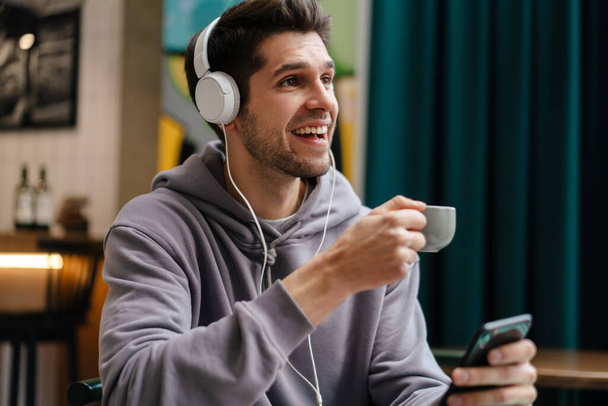 Smiling young casual man in headphones listening to music, holding mobile phone while sitting in cafe indoors, holding cup - Фото, зображення