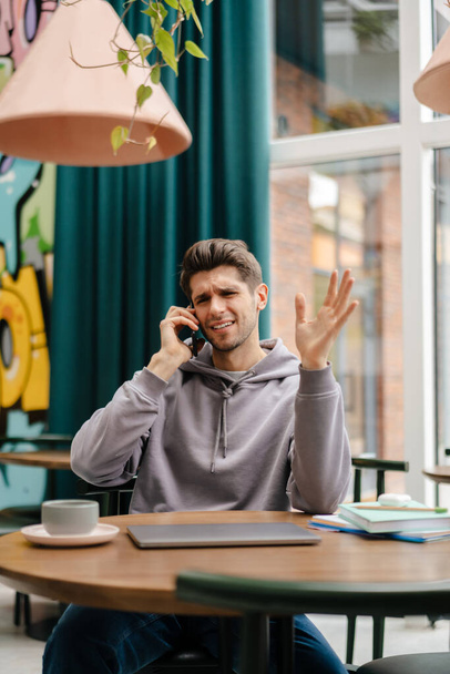 Happy young brunette man holding mobile phone while sitting at the cafe table indoors - Foto, immagini