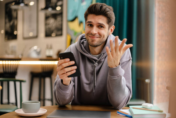 Happy young brunette man holding mobile phone while sitting at the cafe table indoors - Fotografie, Obrázek
