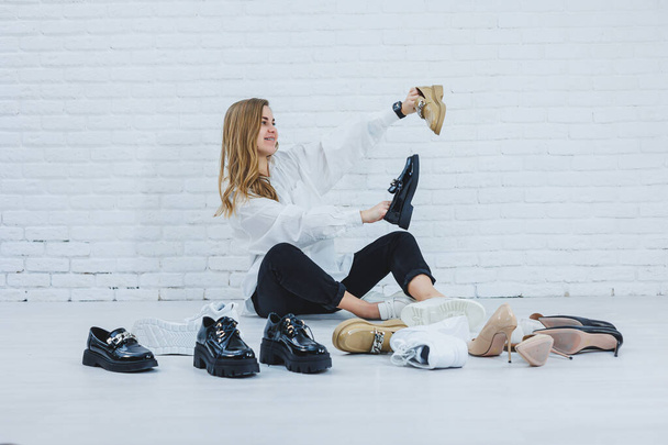 Boldog, gyönyörű, fiatal nő mosollyal, divatos ruhában, fehér ingben és fekete nadrágban ül a padlón a cipők között, és egy új pár cipőt választ magának. - Fotó, kép
