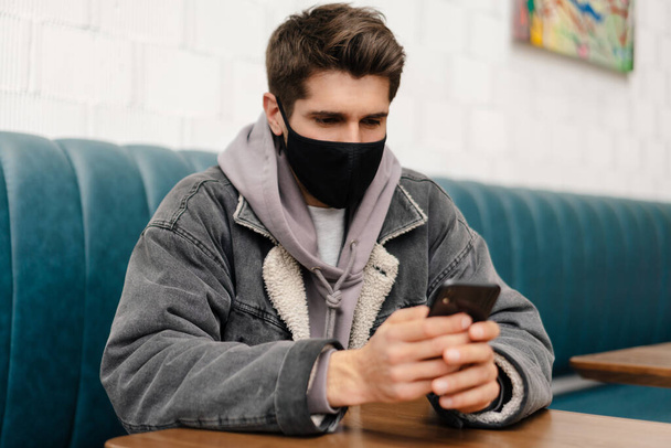 Young brunette man wearing medical mask sitting at the cafe table indoors, holding mobile phone - Photo, Image