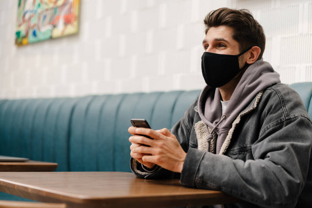 Young brunette man wearing medical mask sitting at the cafe table indoors, holding mobile phone - Foto, Imagen