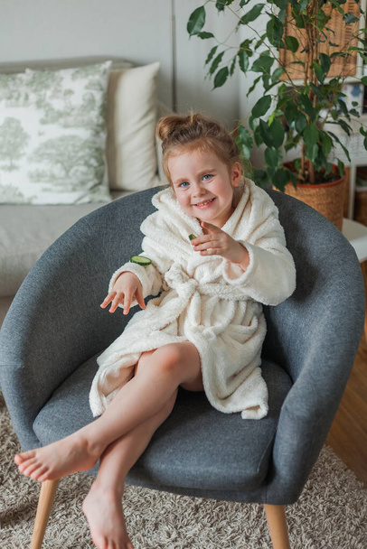 Αξιολάτρευτο κοριτσάκι 3 ετών σε ένα μαλακό φόρεμα σπιτιού με αγγούρια στα χέρια της παίζει στο σαλόνι ομορφιάς στο σπίτι. Το παιδί κάνει θεραπείες ομορφιάς από φυσικά αντικείμενα. - Φωτογραφία, εικόνα