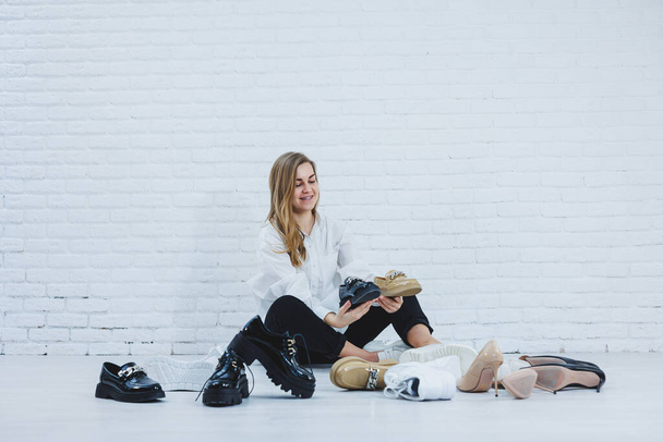Boldog, gyönyörű, fiatal nő mosollyal, divatos ruhában, fehér ingben és fekete nadrágban ül a padlón a cipők között, és egy új pár cipőt választ magának. - Fotó, kép