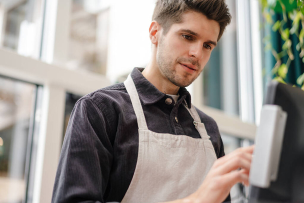Serious young man in apron standing at the cash register indoors - Foto, imagen