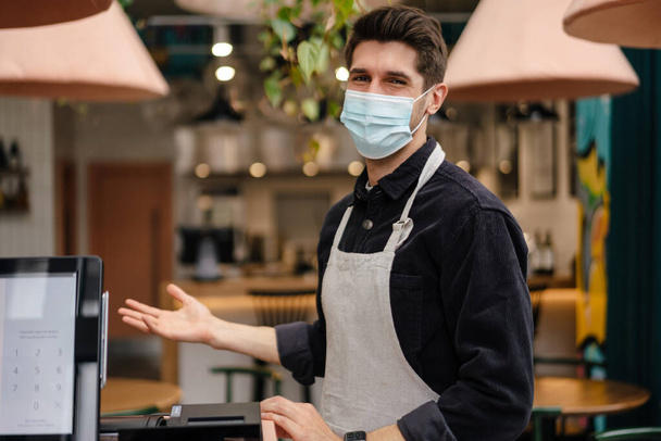 Young man in apron wearing protective medical mask standing at the cash register in coffee shop - Zdjęcie, obraz