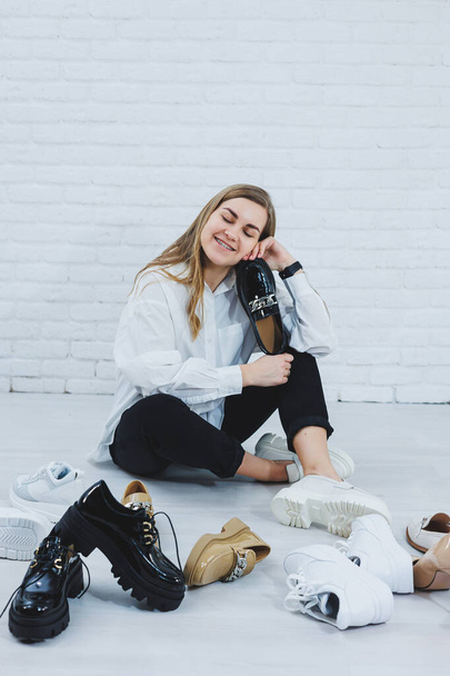 Mladá žena sedí na podlaze mezi botami a vybírá si nové boty pro sebe. Dáma v bílé košili a černých kalhotách. Žena v bílých botách. - Fotografie, Obrázek