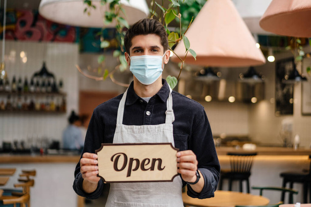 Young man waiter in apron showing open sign while standing indoors the cafe - 写真・画像