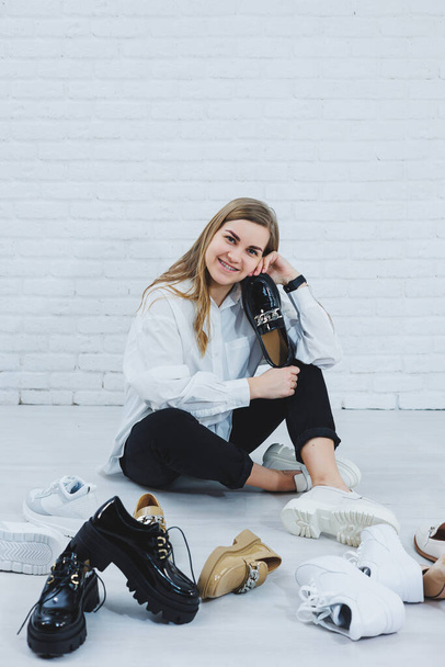 A young woman sits on the floor among shoes and chooses new shoes for herself. Lady in a white shirt and black trousers. Woman in white shoes. - Photo, Image