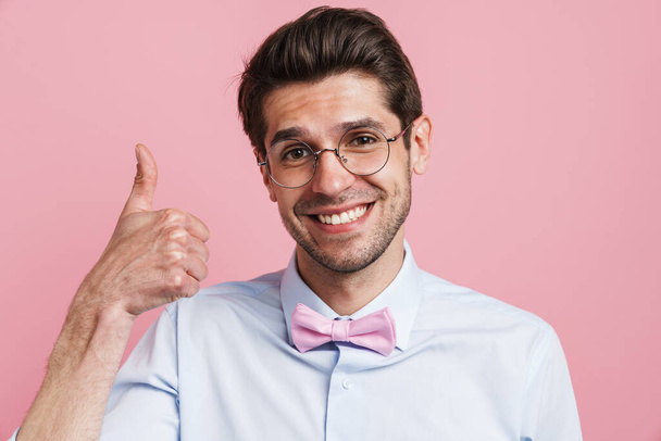 Portrait of a smiling young brunette nerd man wearing shirt and bowtie standing over pink wall background showing thumbs up - Valokuva, kuva