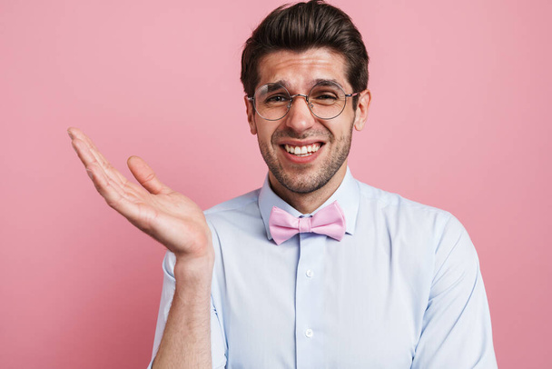 Portrait of a confused frowning young brunette nerd man wearing shirt and bowtie standing over pink wall background - Photo, Image