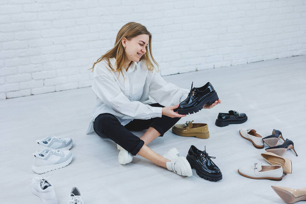 Una mujer se sienta en el suelo en un vestidor entre zapatos y elige zapatos nuevos para ella. Dama con camisa blanca. Mujer en zapatos blancos. - Foto, imagen