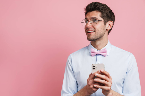 Young white man wearing bow tie smiling and using cellphone isolated over pink background - 写真・画像