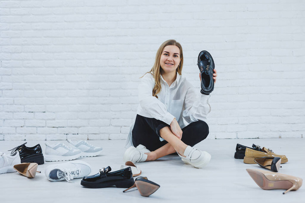 A young woman sits on the floor among shoes and chooses new shoes for herself. Lady in a white shirt and black trousers. Woman in white shoes. - Foto, afbeelding