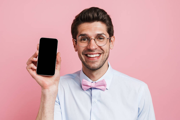 Young white man wearing bow tie smiling and showing cellphone isolated over pink background - Fotografie, Obrázek