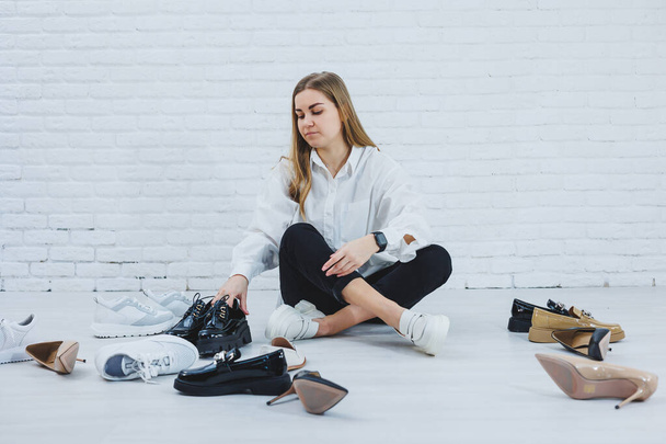 Elegante rubia eligiendo zapatos mientras está sentada en el suelo. Vista lateral de una gran cantidad de zapatos de mujer en el suelo, una selección de zapatos elegantes. - Foto, imagen