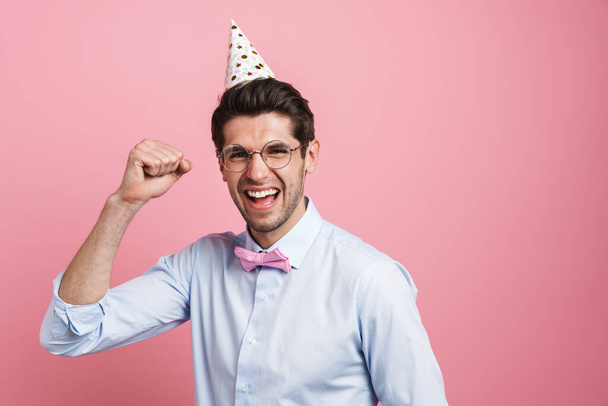 Young white man wearing party cone laughing and making winner gesture isolated over pink background - Zdjęcie, obraz