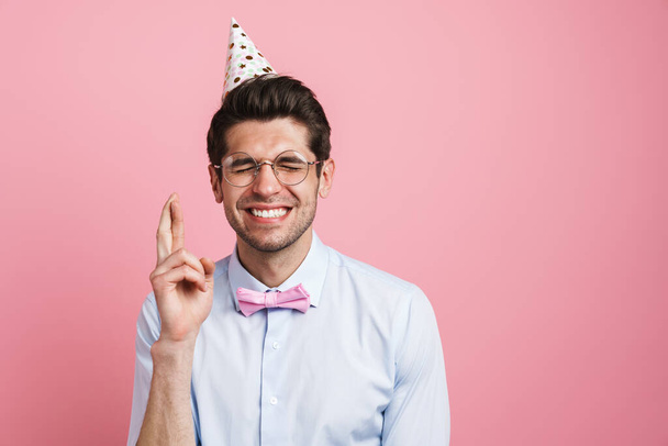 Young man wearing bow tie and party cone holding fingers crossed isolated over pink background - Photo, Image