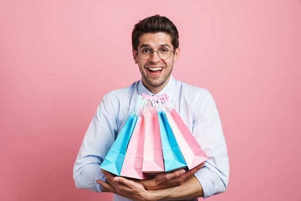 Young white man wearing bow tie smiling while posing with shopping bags isolated over pink background - Fotoğraf, Görsel