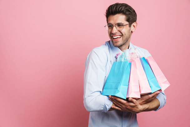 Young white man wearing bow tie smiling while posing with shopping bags isolated over pink background - Foto, afbeelding