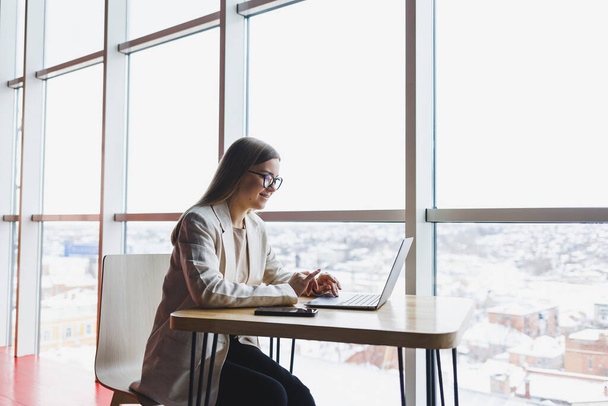 Mulher focada em roupas casuais digitando em um netbook enquanto trabalhava em um novo projeto, sentada em uma mesa em um espaço de trabalho moderno. Mulher de negócios de casaco e óculos. Trabalho remoto - Foto, Imagem