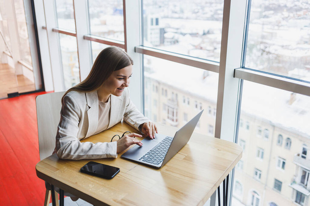 Összpontosított nő alkalmi viseletben gépel egy netbookon, miközben egy új projekten dolgozik, egy íróasztalnál ül egy modern munkahelyen. Üzletasszony kabátban és szemüvegben. Távoli munka - Fotó, kép