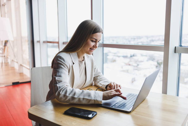 Modern bir ofiste geniş pencereli bir ofiste telefonla konuşurken ceketli, gülümseyen ve dizüstü bilgisayarla çalışan mutlu genç bir kadının görüntüsü. Uzak çalışma - Fotoğraf, Görsel