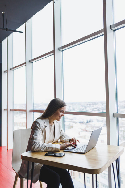 Imagen de una joven feliz con una chaqueta sonriendo y trabajando en un portátil mientras habla por teléfono en una oficina moderna con grandes ventanales. Trabajo remoto - Foto, imagen