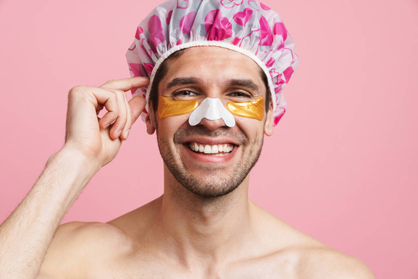 Young shirtless man with eye and nose patches smiling at camera isolated over pink background - Zdjęcie, obraz
