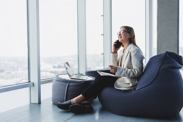 Business woman thinks while writing in a notebook at work. Modern successful woman. Young smiling girl in glasses sits in a chair at a table in an open office - Fotografie, Obrázek